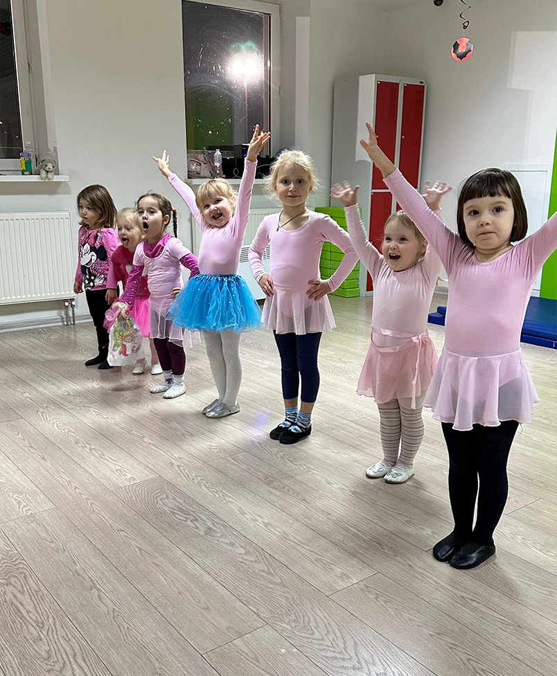 Група маленьких дівчаток у рожевому балетному одязі стоїть у ряд з піднятими руками.